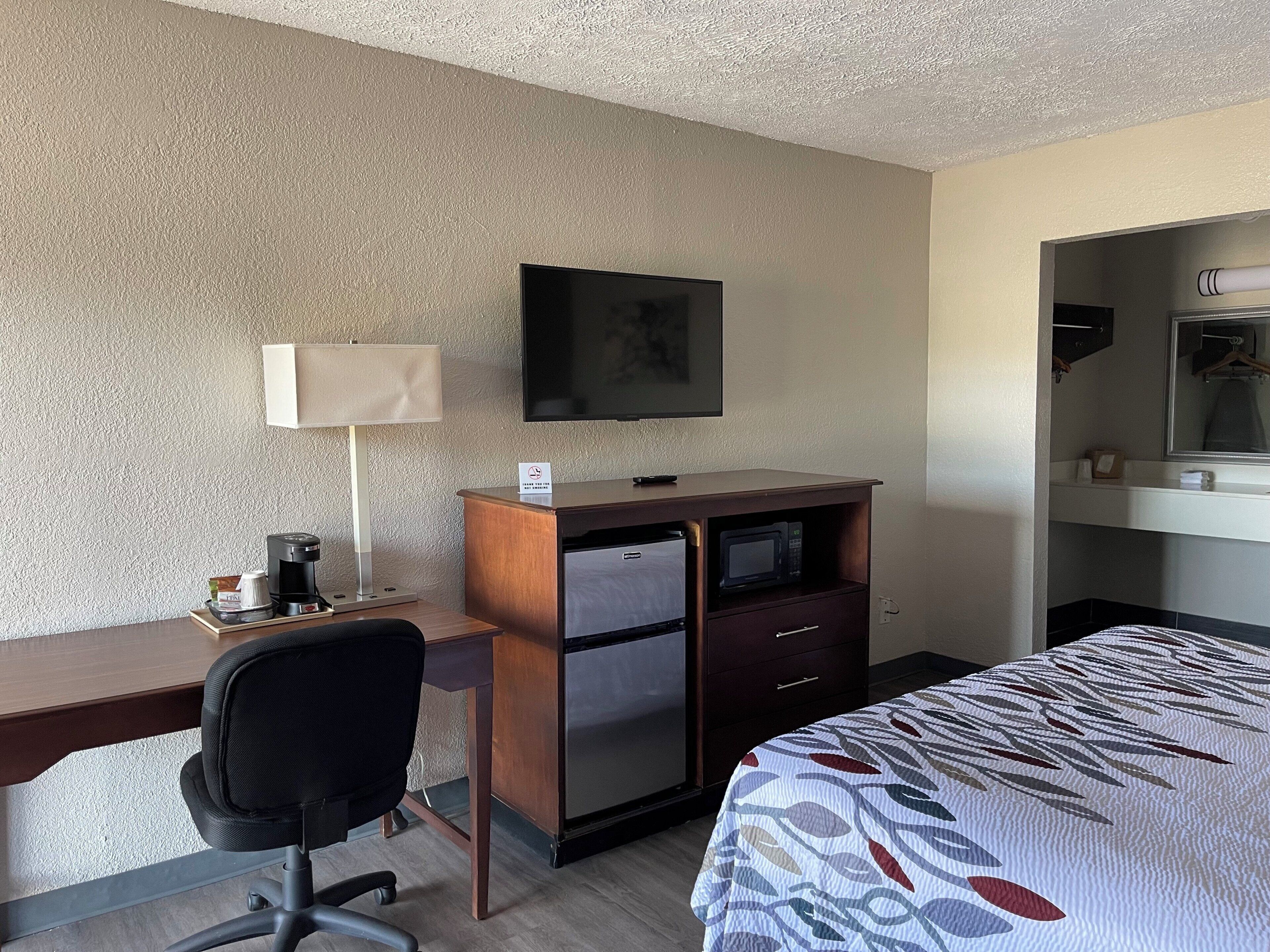 A hotel room with a desk, a chair, a bed, a television, a refrigerator, and a microwave.