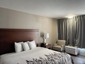 A hotel room with a large bed, a bedside table, a lamp, a chair, and a window with curtains. - Red Roof Inn LaGrange (LaGrange)