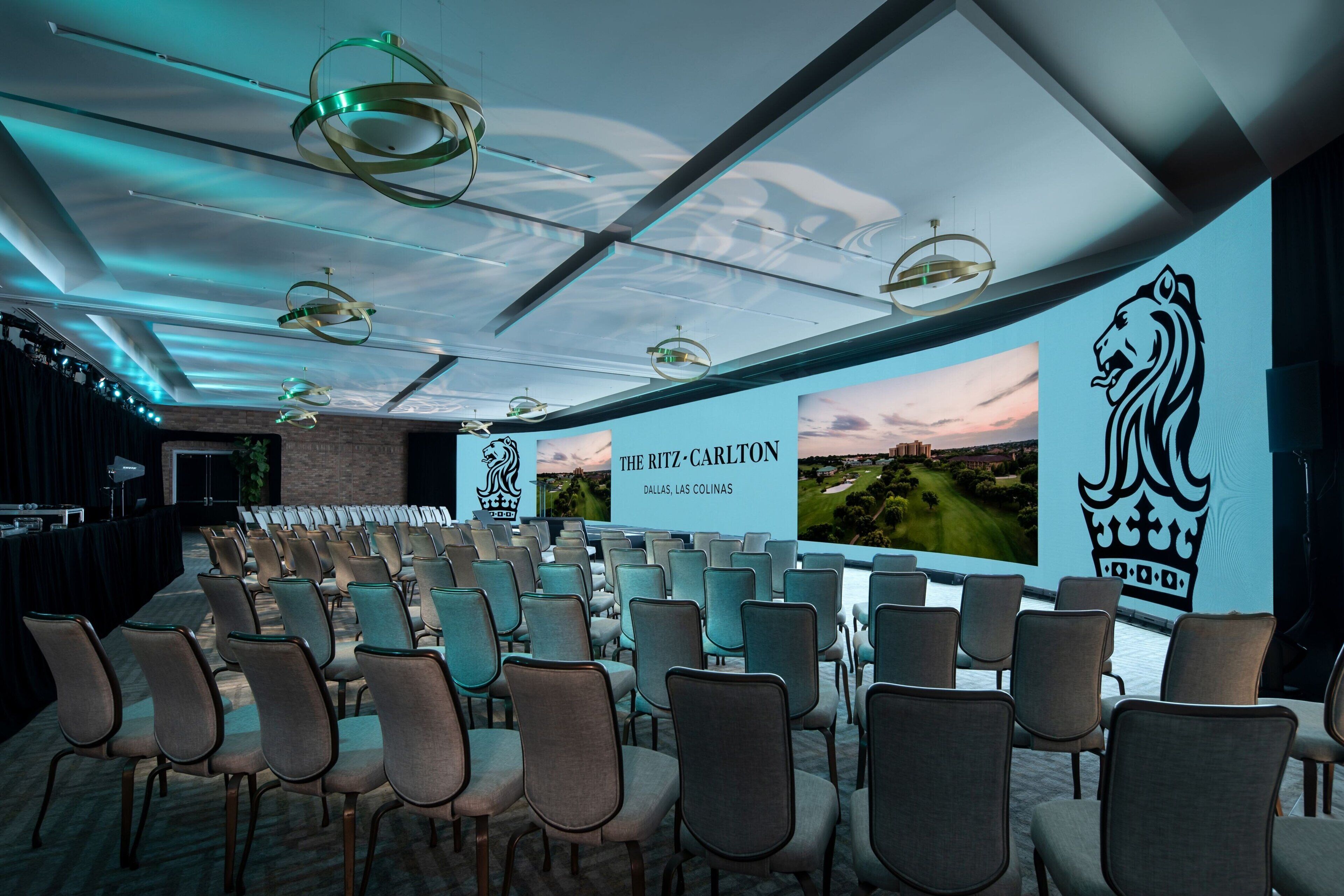 A conference room with rows of chairs, a large screen displaying 'The Ritz-Carlton Dallas, Las Colinas', and a lion logo on the wall.