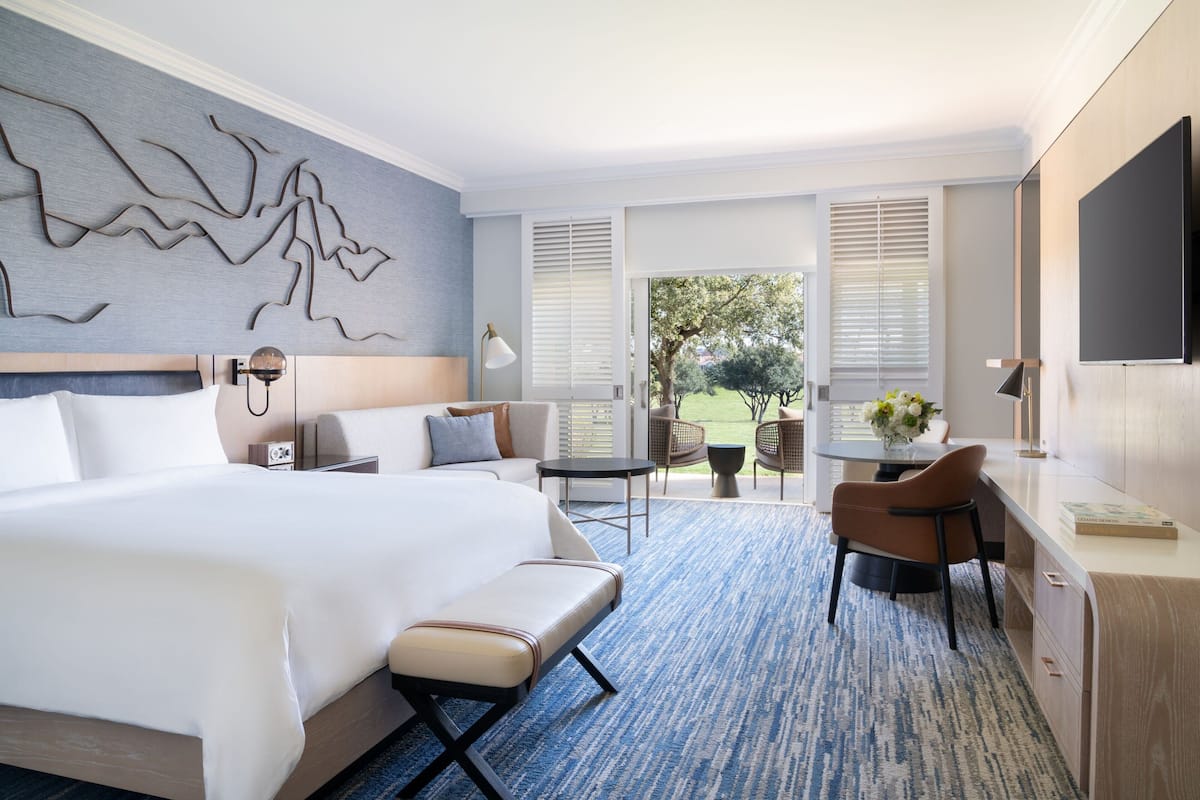 Habitación de hotel con una cama grande, un escritorio, una silla, una mesita y un televisor. Hay una ventana con persianas, una obra de arte en la pared y vistas al exterior.