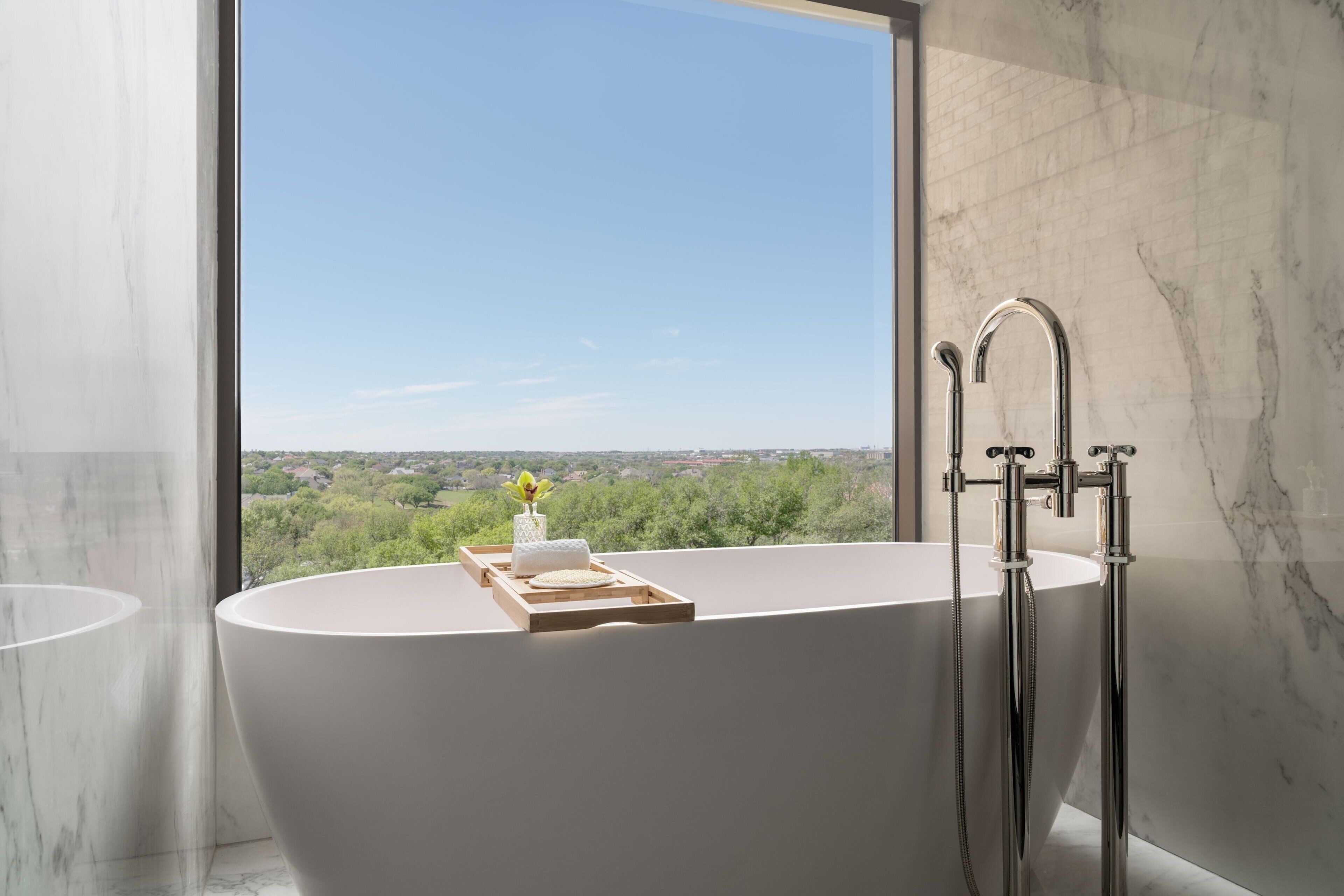 A modern bathroom with a freestanding bathtub, a large window offering a scenic view, and marble walls.
