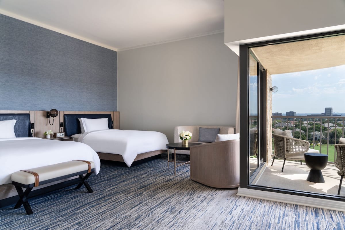 Habitación de hotel con dos camas, un banco, una mesita con un florero y un balcón con sillas de mimbre.