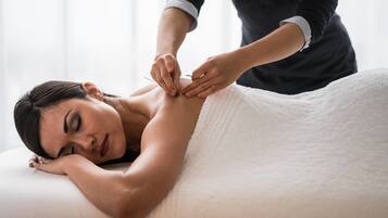 A person receiving a massage on a white towel-covered bed in a room with a plain white wall.