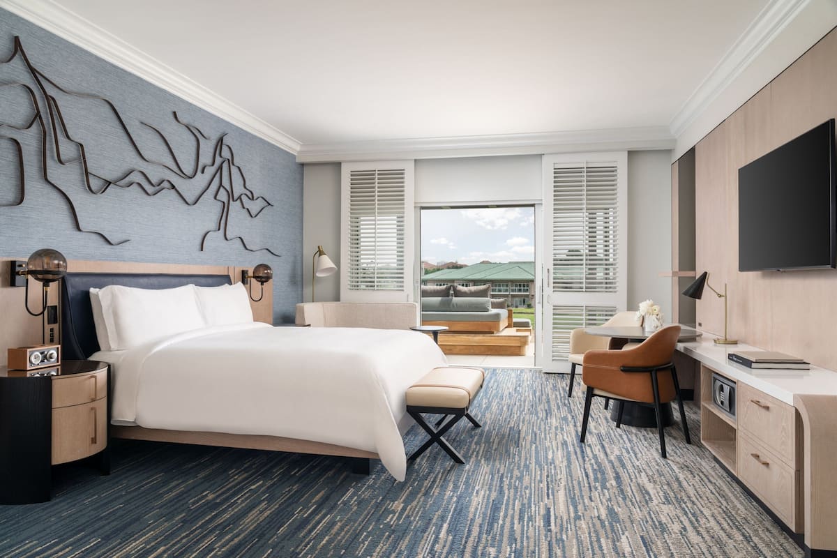 Una habitación de hotel moderna con una cama grande, un escritorio, una silla y un televisor. Se ve el exterior a través de las ventanas.