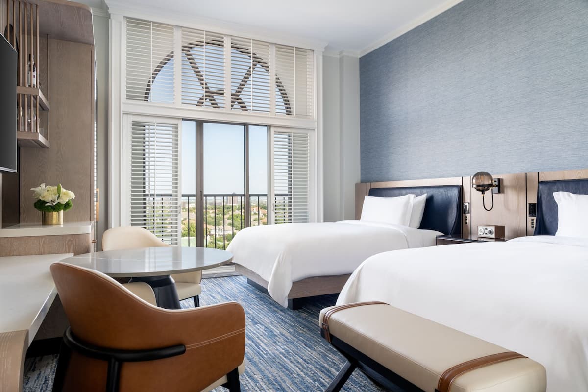 Habitación de hotel con dos camas, un escritorio, una silla y vistas al exterior a través de amplios ventanales.