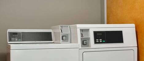 Deux appareils électroménagers blancs, probablement une machine à laver et un sèche-linge, sont placés l’un sur l’autre dans une pièce dotée d’une porte en bois et d’un mur sobre.