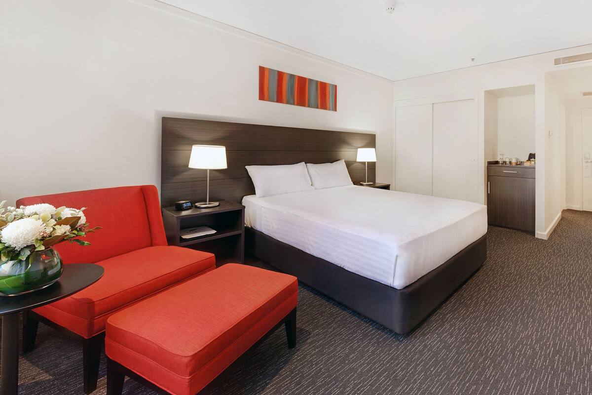 Quarto de hotel com cama, escrivaninha, cadeira, mesa pequena com um vaso de flores e um banco vermelho.