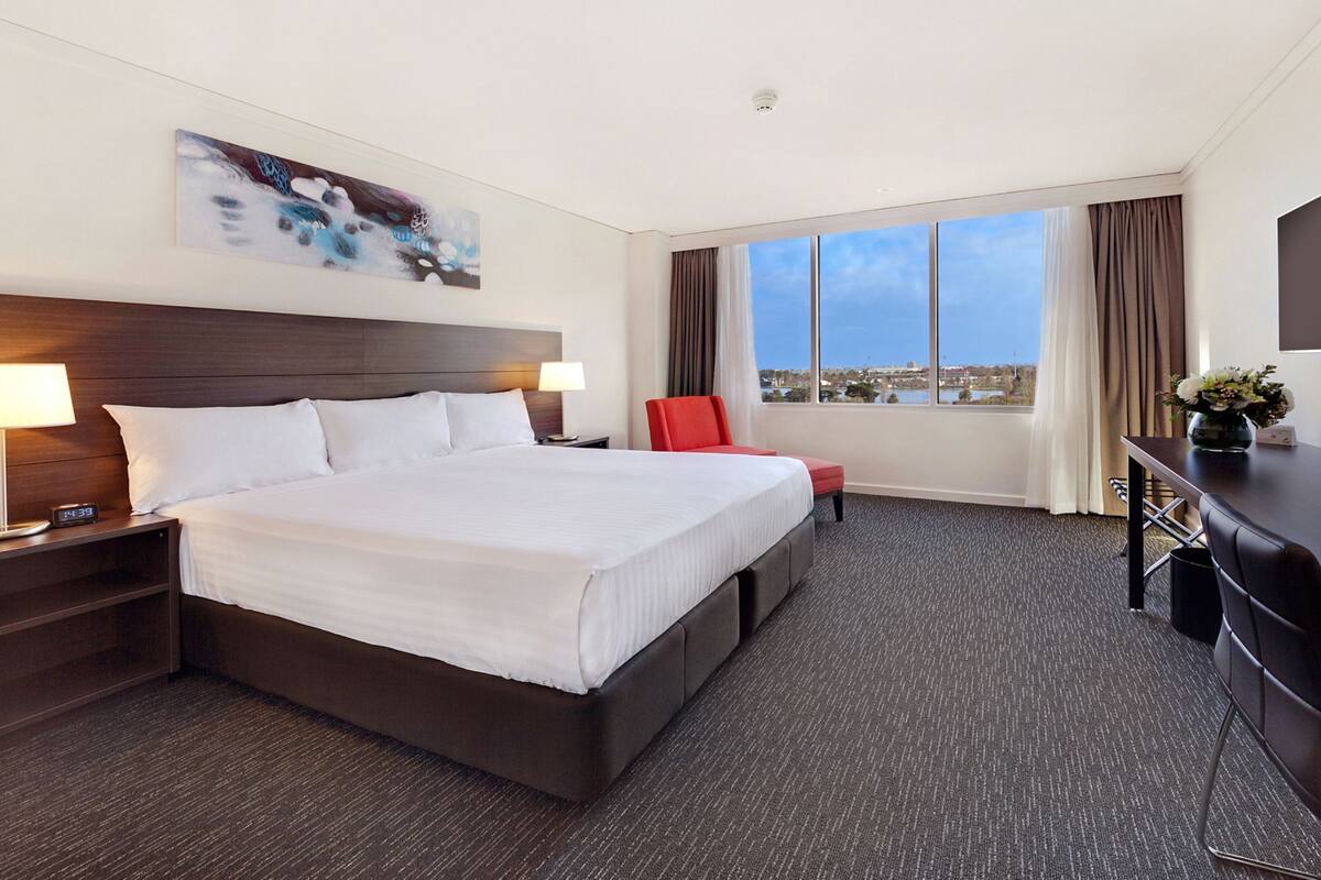 Quarto de hotel com uma cama grande, duas luminárias de cabeceira, uma escrivaninha com cadeira, uma televisão de tela plana e uma janela com vista para a cidade.
