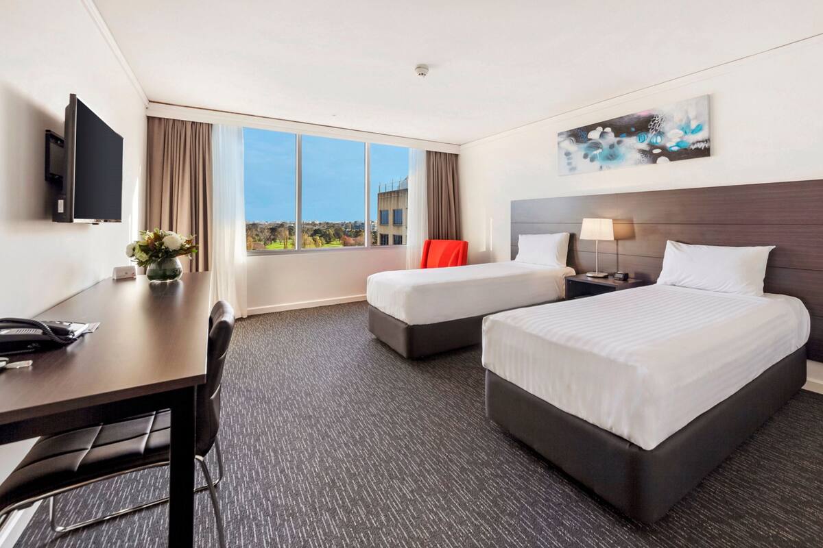 Quarto de hotel com uma cama grande, uma cama menor, uma escrivaninha com telefone, televisão e janela com vista para edifícios e árvores.