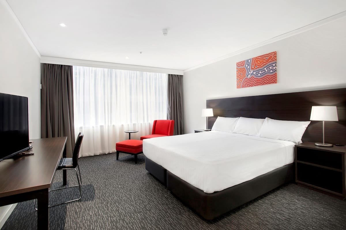 Quarto de hotel com uma cama grande, uma escrivaninha com televisão, uma cadeira vermelha e uma janela com cortinas.