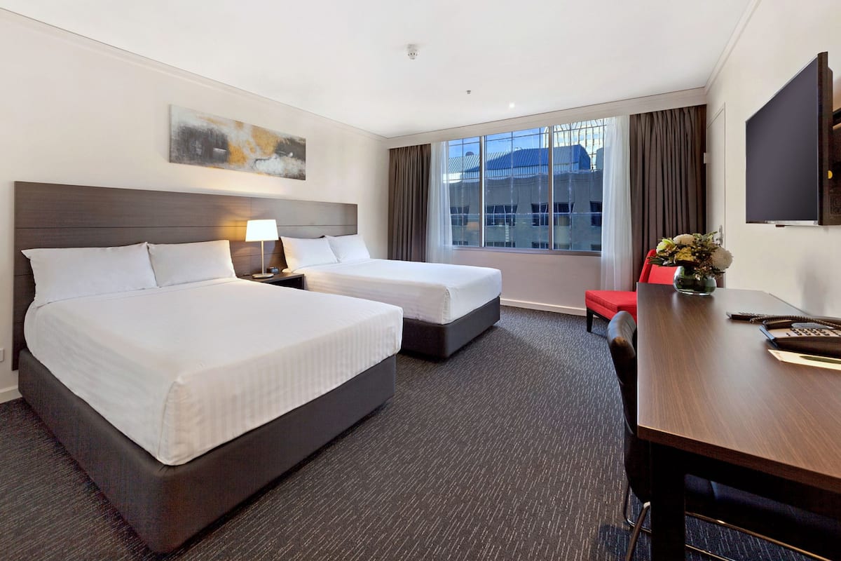 Quarto de hotel com duas camas, uma escrivaninha, televisão e janela com vista para edifícios.