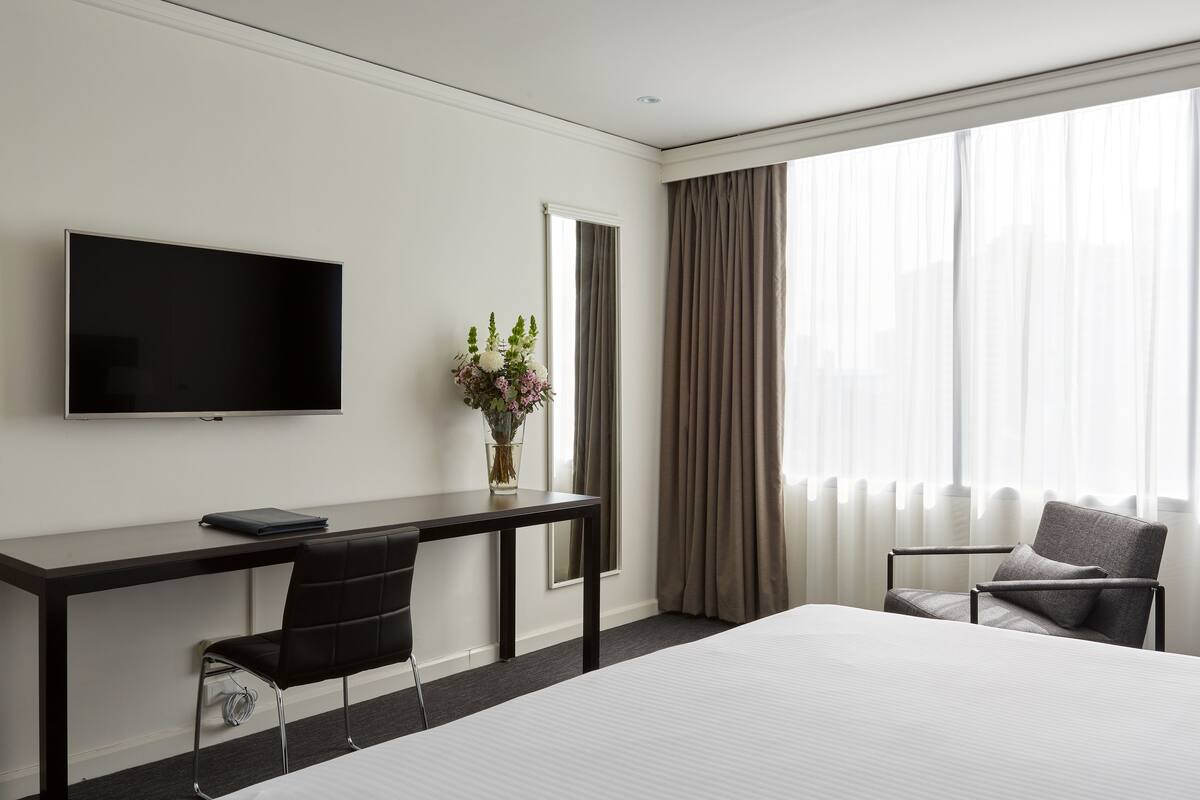 Quarto de hotel com cama, secretária com cadeira, televisão, um vaso com flores e janela com cortinas.
