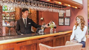 Et barområde med en bartender og en gæst. Der hænger vinglas over baren, en vinflaske står på disken, og der er en menu på væggen.