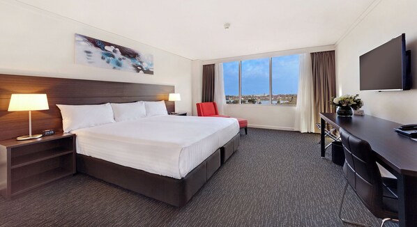 A hotel room with a large bed, a desk, a chair, a TV, and a window with a city view.