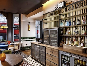 A bar area with a variety of liquor bottles, a refrigerator, and a seating area with a view of a train outside. - ibis Styles London Gloucester Road (London)