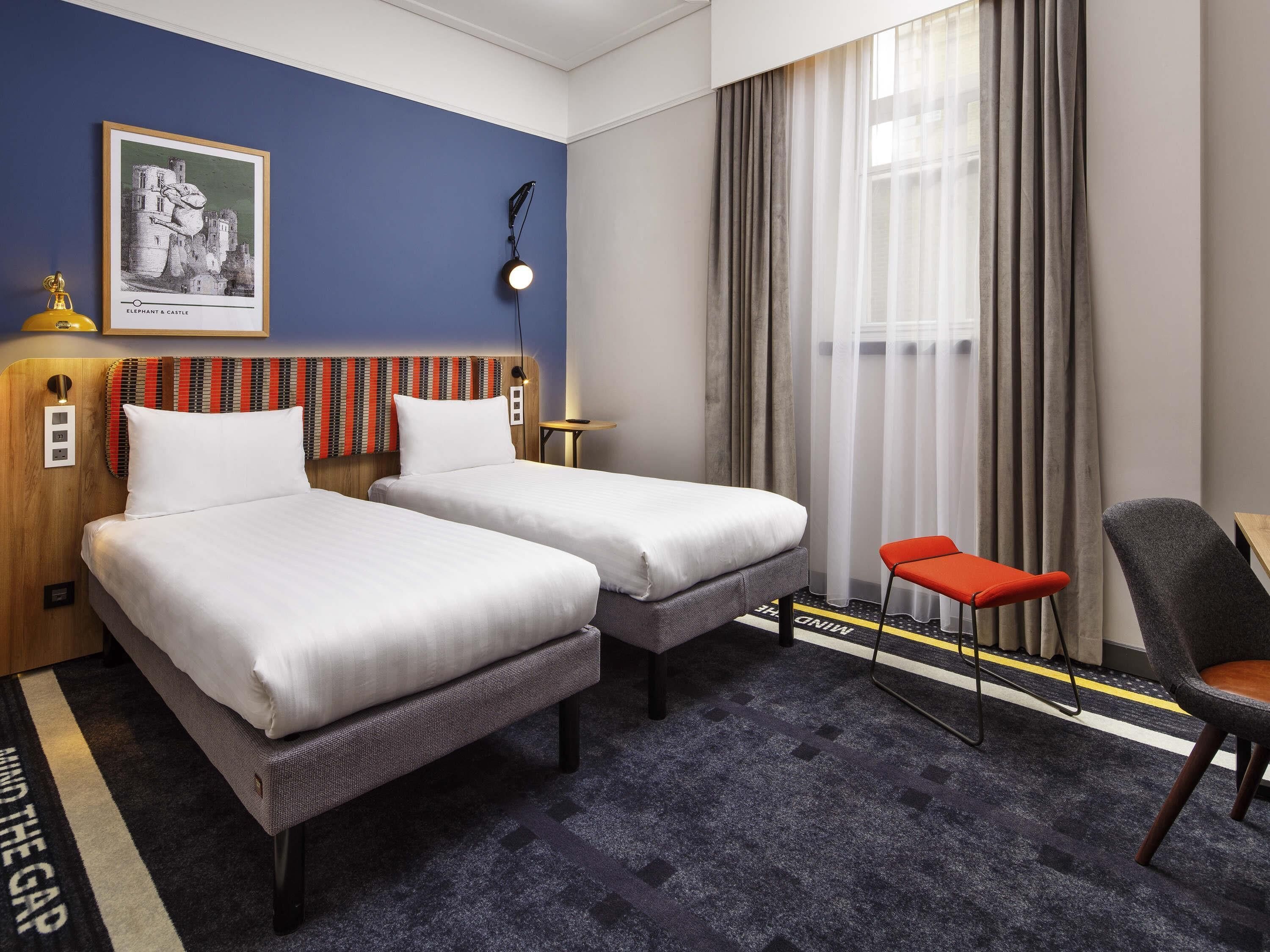 A hotel room with two beds, a desk, a chair, a lamp, a painting, and a window with curtains.