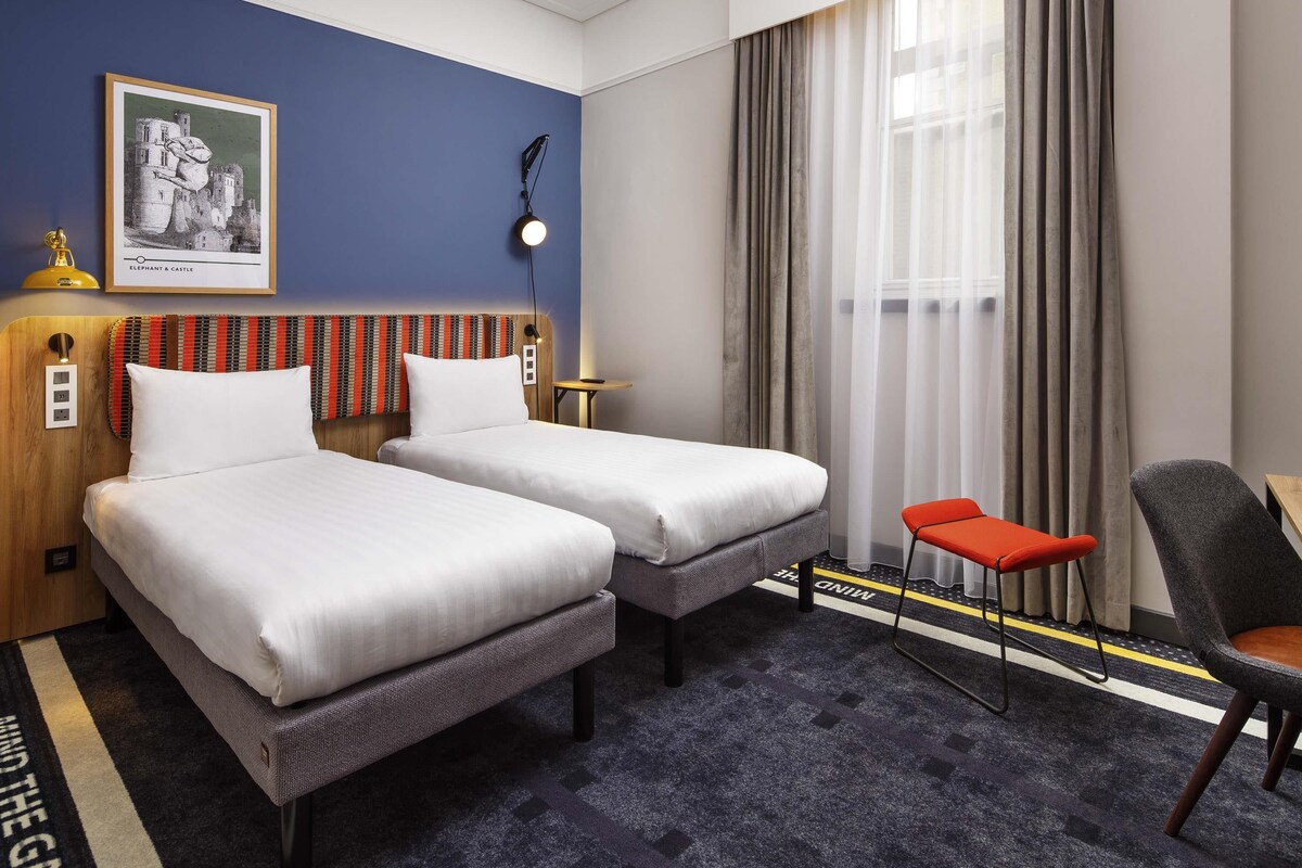 A hotel room with two beds, a desk, a chair, a lamp, a painting, and a window with curtains.
