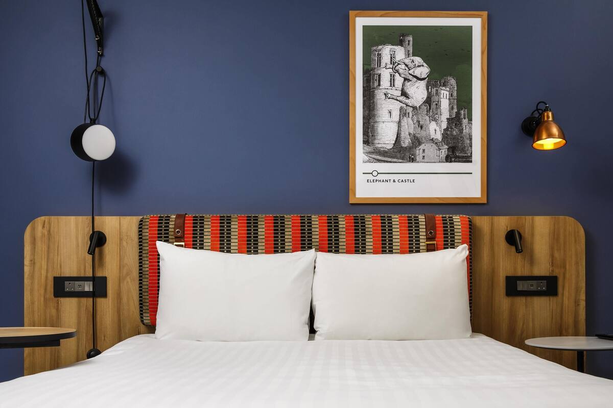 A hotel room with a wooden headboard, two white pillows, a framed picture of an elephant and castle, a bedside lamp, and a small table with a power outlet.