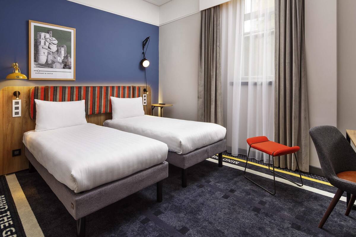 Una habitación de hotel con dos camas, un escritorio, una silla, una lámpara, un cuadro y una ventana con cortinas.
