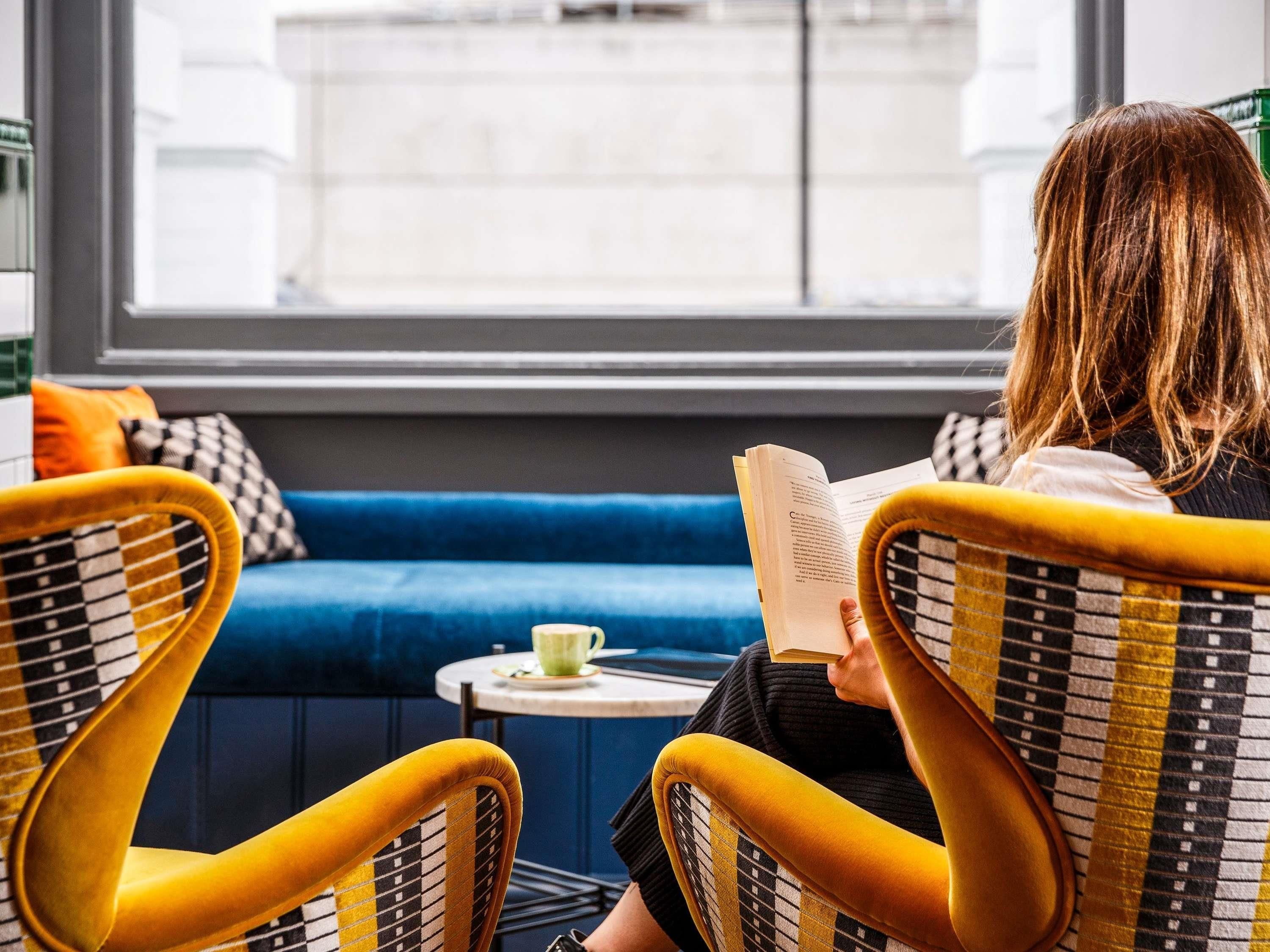 A person seated on a yellow chair reading a book at a small table with a cup, in a room with a blue sofa and a window with a view of a building.