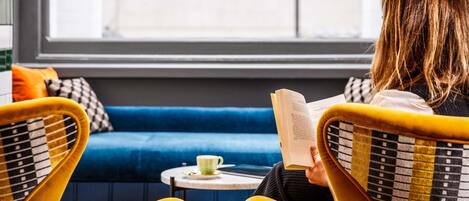 Una persona sentada en una silla amarilla leyendo un libro en una mesa pequeña con una taza, en una habitación con un sofá azul y una ventana que ofrece vistas a un edificio.