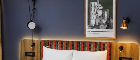 A hotel room with a bed, two bedside tables, a wall-mounted lamp, a framed picture, and a small round table.