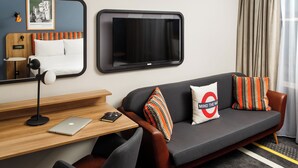 A modern hotel room with a grey sofa, wooden desk, and a large flat-screen TV.