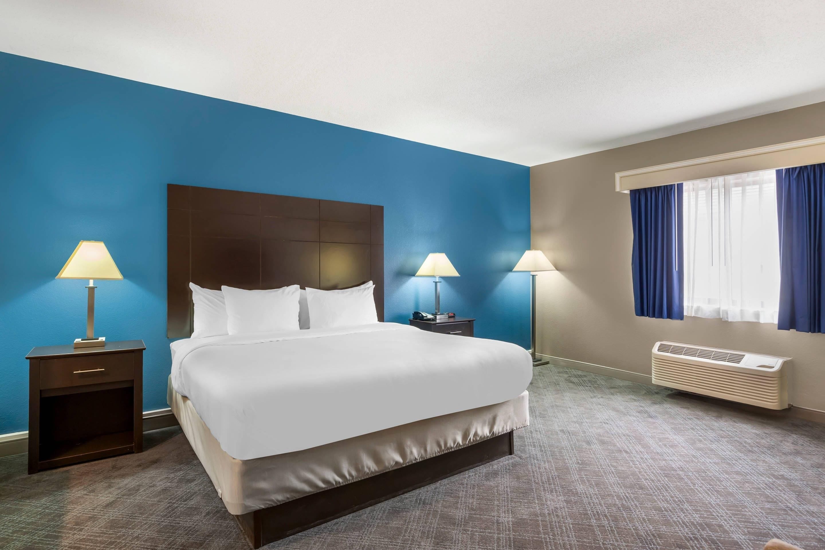 Een hotelkamer met een groot bed, twee nachtkastjes met lampen, een raam met gordijnen en een wandairconditioner.