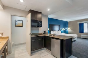 A compact hotel room with a kitchenette, a bed, a desk, and a bathroom.