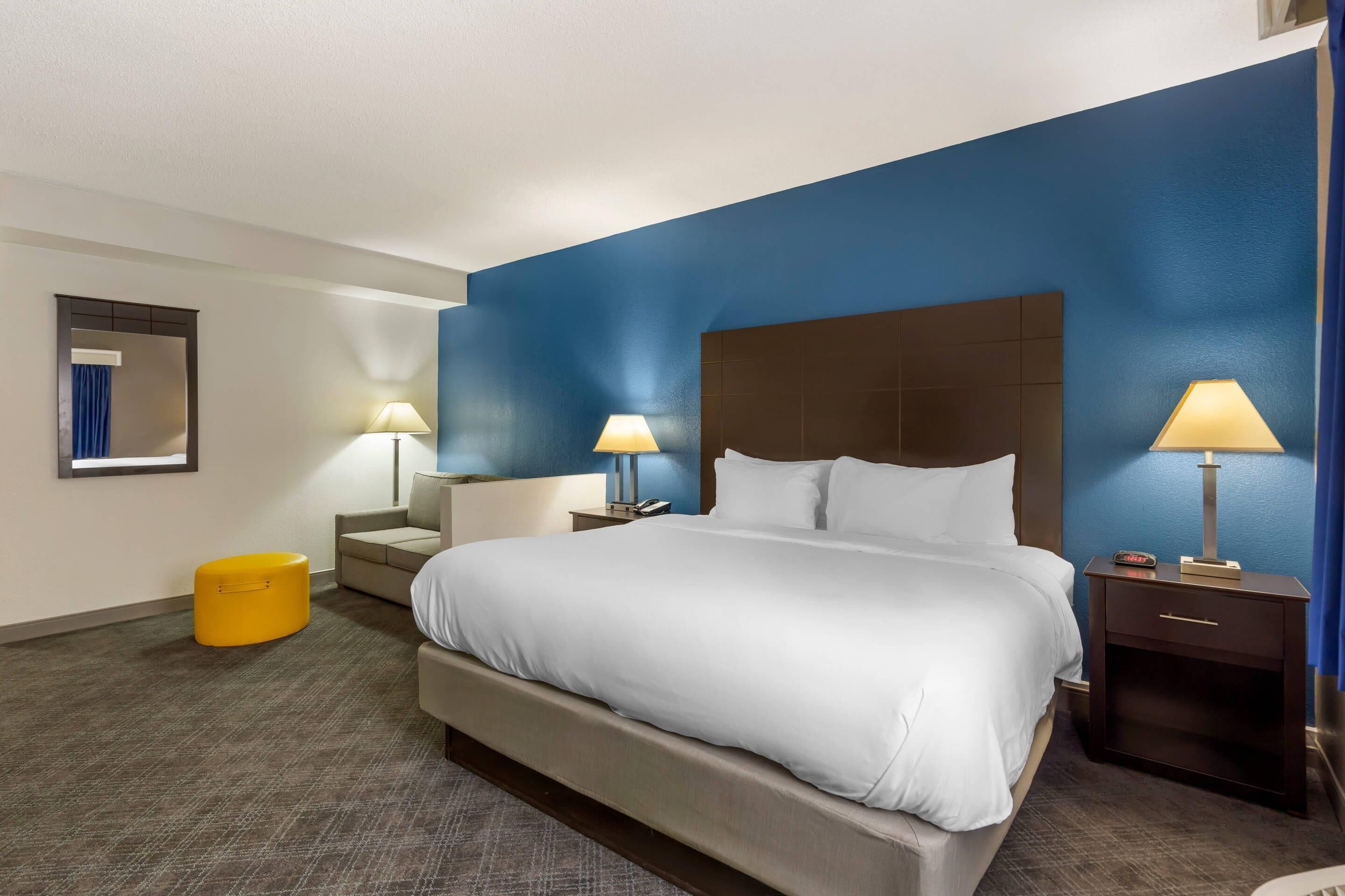 Una habitación de hotel con una cama grande, dos mesitas de noche con lámparas, un pequeño taburete amarillo y un espejo en la pared.