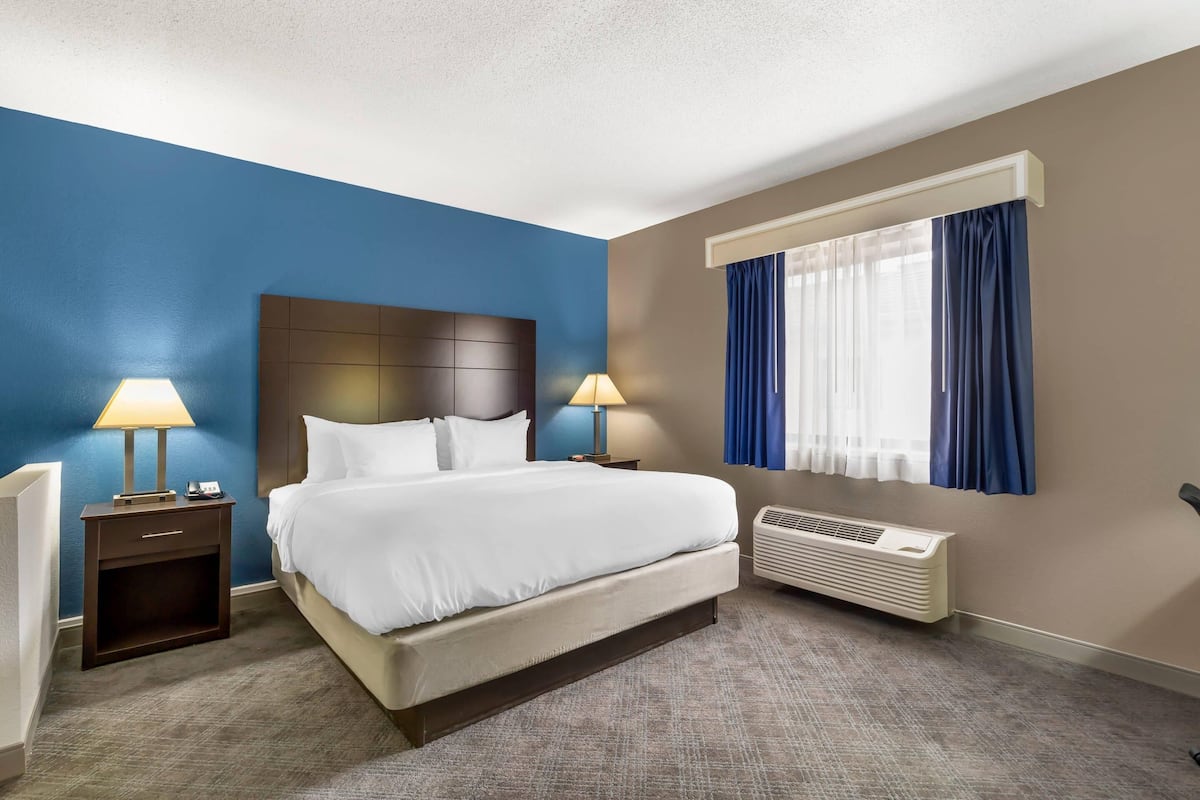 A hotel room with a large bed, two bedside tables with lamps, a window with curtains, and a wall-mounted air conditioner.