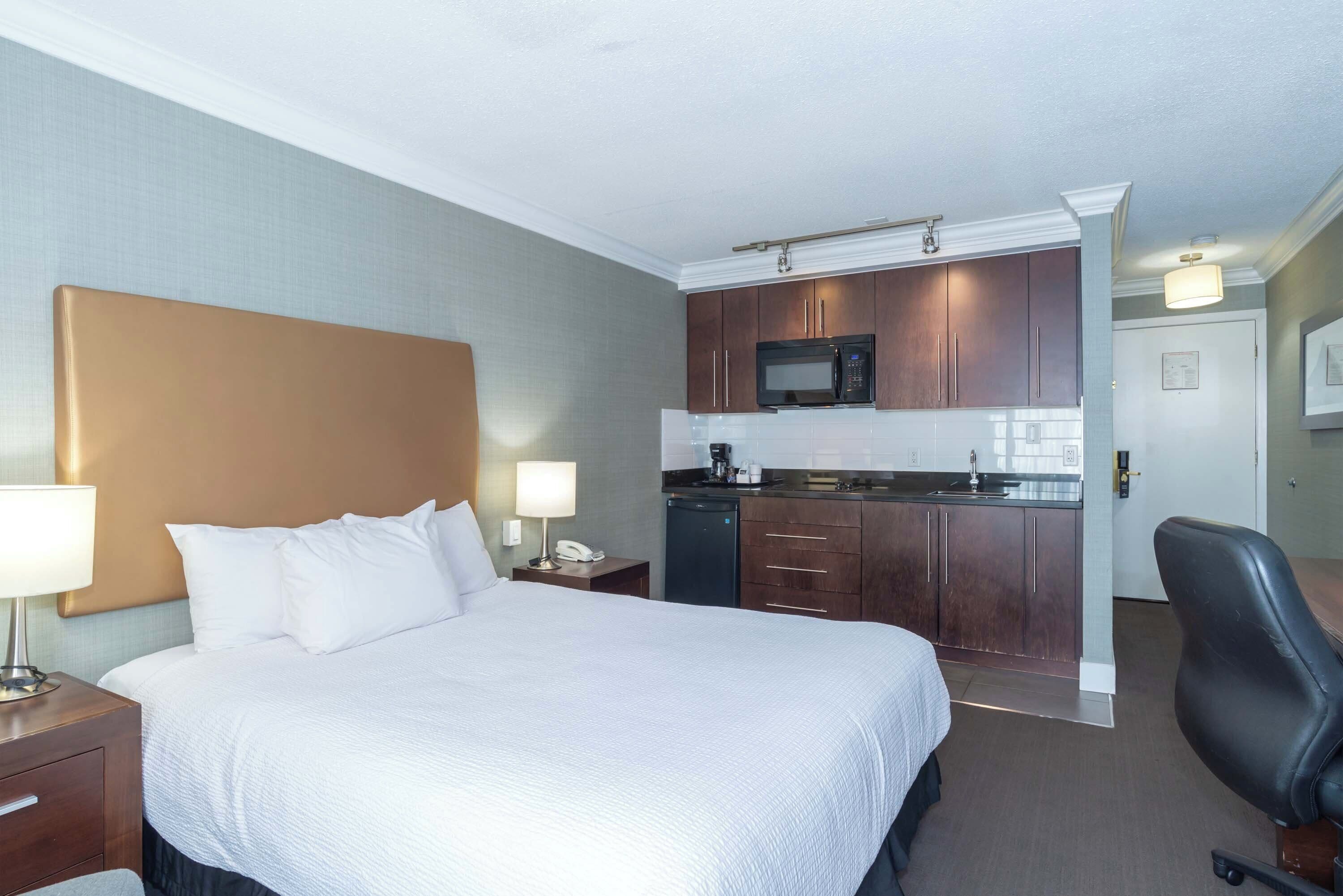 Une chambre d’hôtel comprenant un lit, des lampes de chevet, un bureau avec une lampe, une chaise, une kitchenette équipée d’un four à micro-ondes et un petit réfrigérateur.