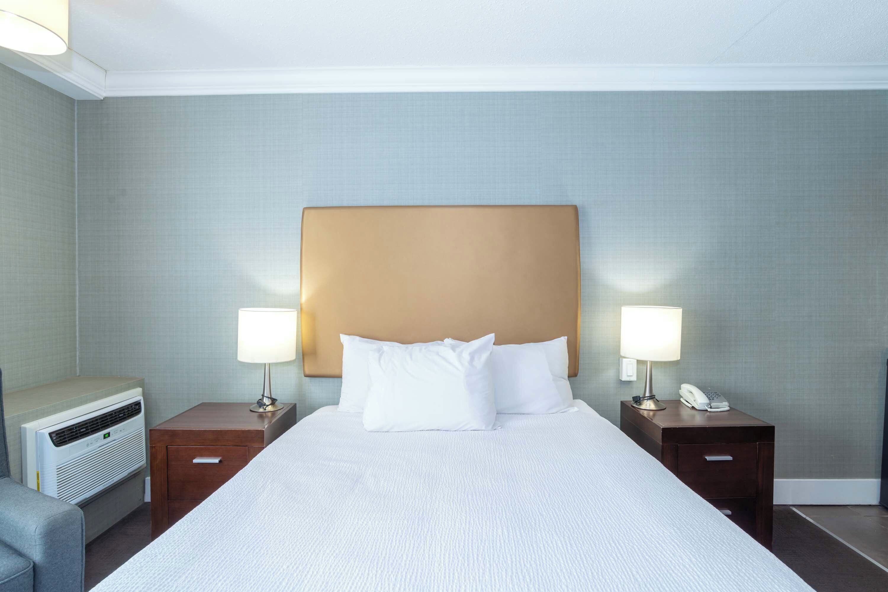 Una habitación de hotel con una cama grande, dos mesitas de noche con lámparas, un calentador y un sillón pequeño.