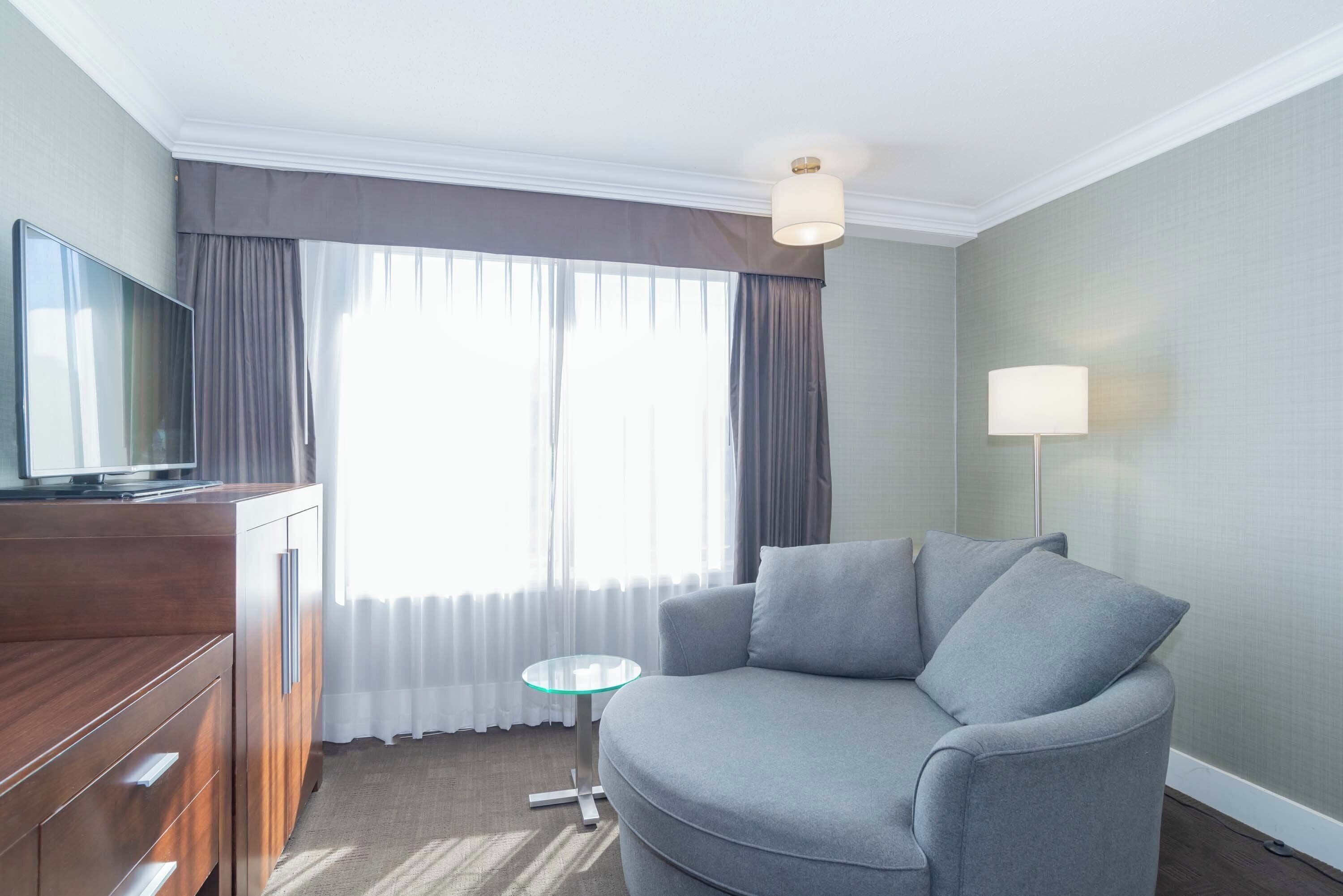 A modern hotel room with a grey sofa, a wooden cabinet with a flat-screen TV, a small glass table, and a lamp.