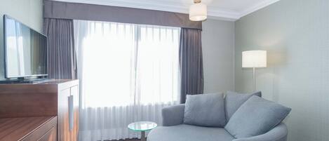 A modern hotel room with a grey sofa, a wooden cabinet with a flat-screen TV, a small glass table, and a lamp.