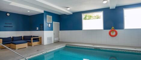 An indoor swimming pool with blue walls, a white tiled floor, and a seating area with cushions.