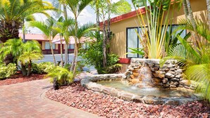 Un jardín ajardinado con una fuente de piedra, palmeras y un edificio de techo rojo.