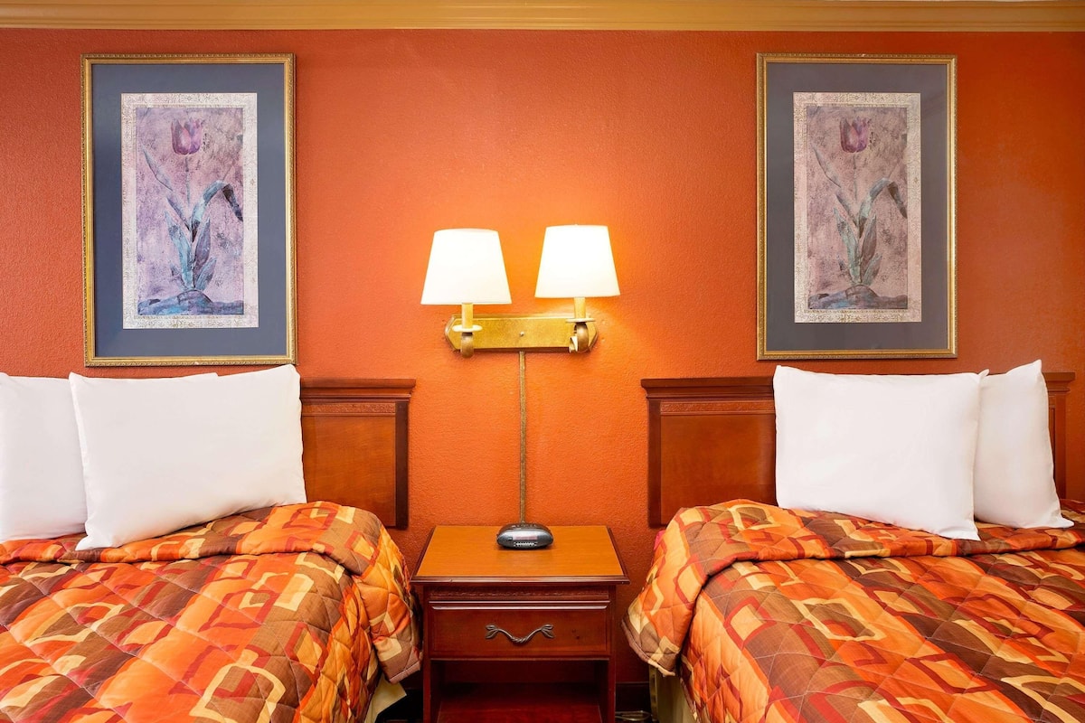 A hotel room with two beds, a nightstand with a lamp, and two framed floral paintings on the walls.