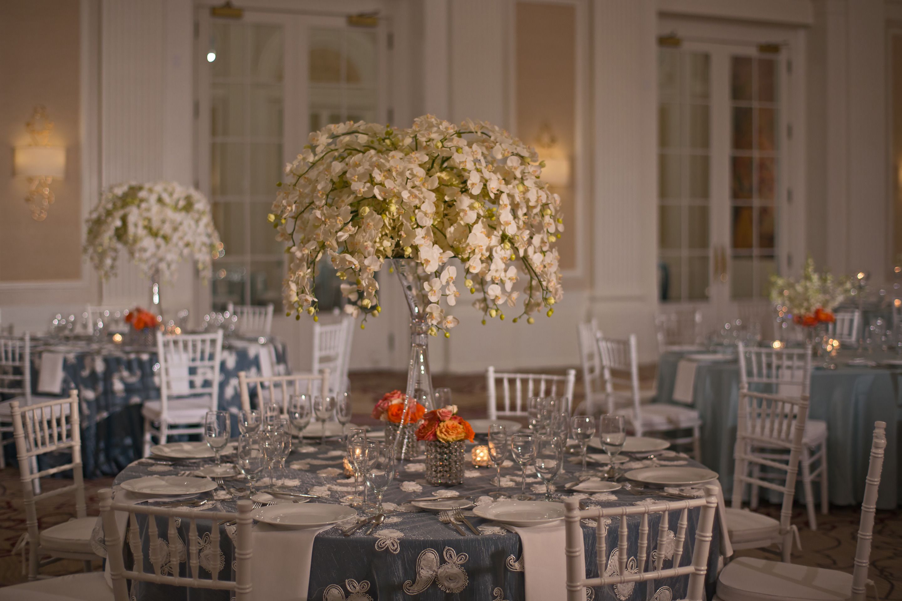 Un salón de banquetes con mesas redondas dispuestas para comer, cada una con un centro de mesa floral y sillas blancas.