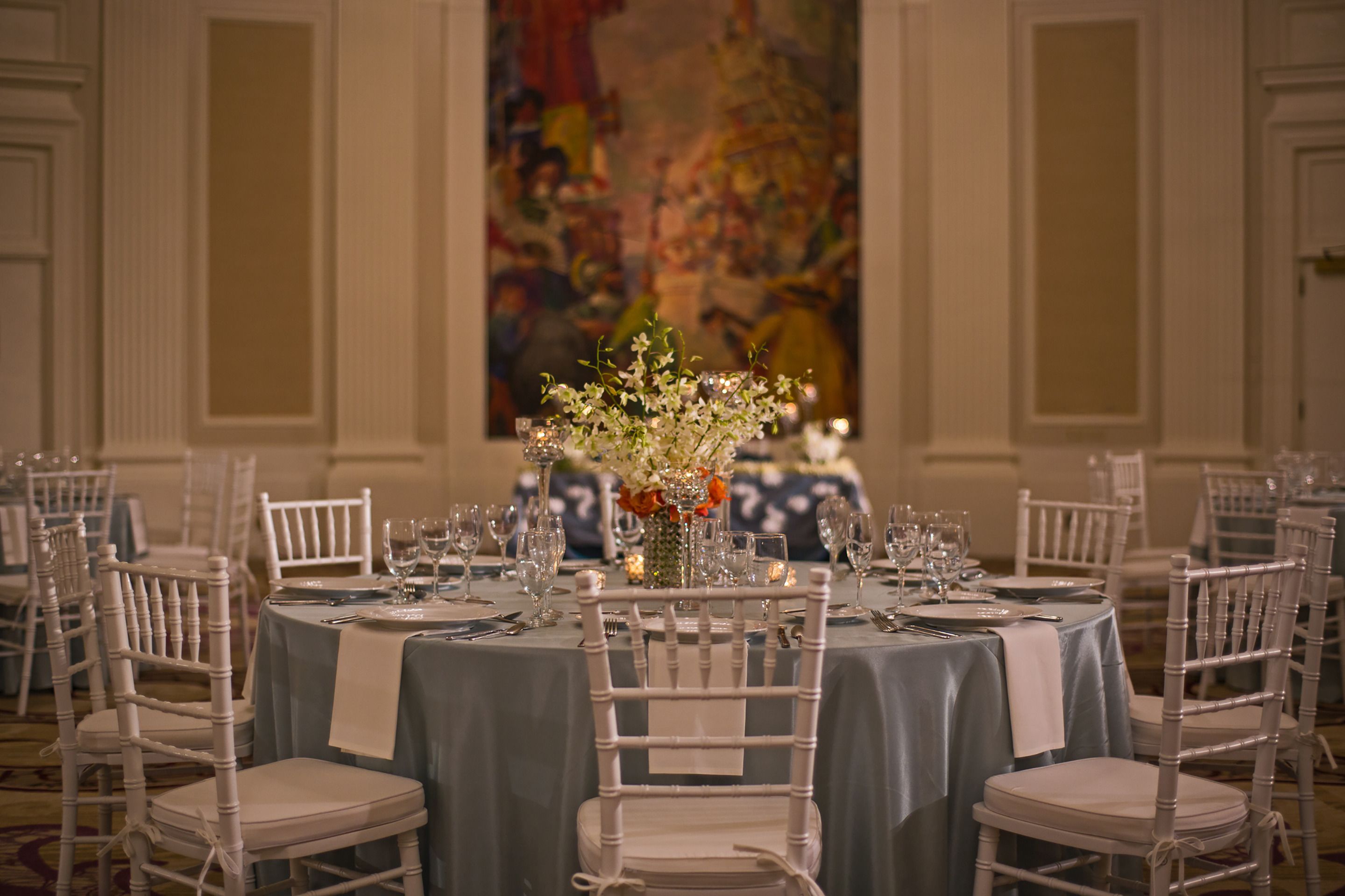 A formal dining room with round tables, white chairs, and a large floral centerpiece.