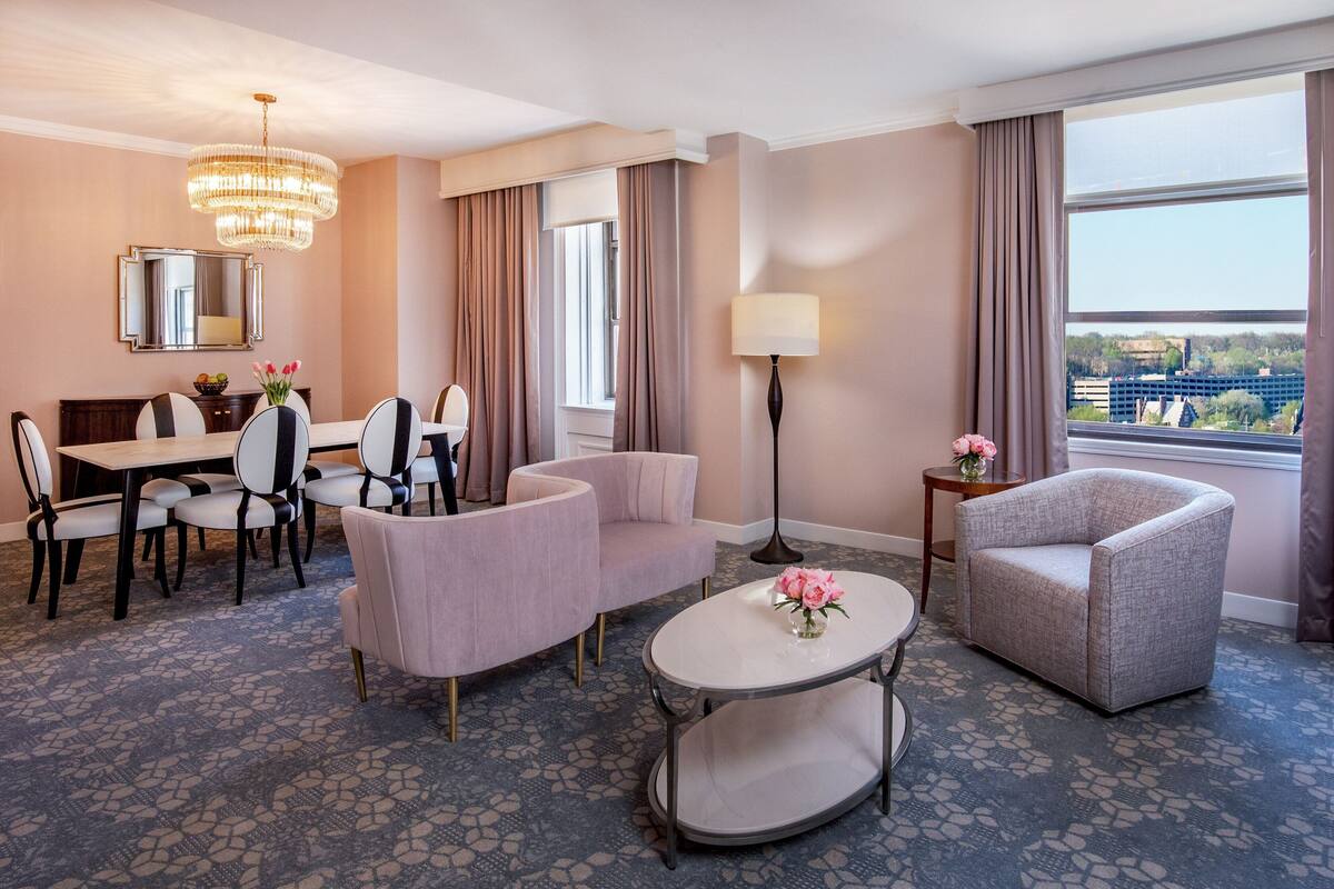 Hotelkamer met een zithoek, twee fauteuils, een ronde tafel, een vloerlamp en een groot raam met uitzicht over de stad.