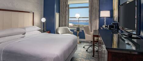 A hotel room with a large bed, a desk with a chair, a TV, and a view of the sea through the window.