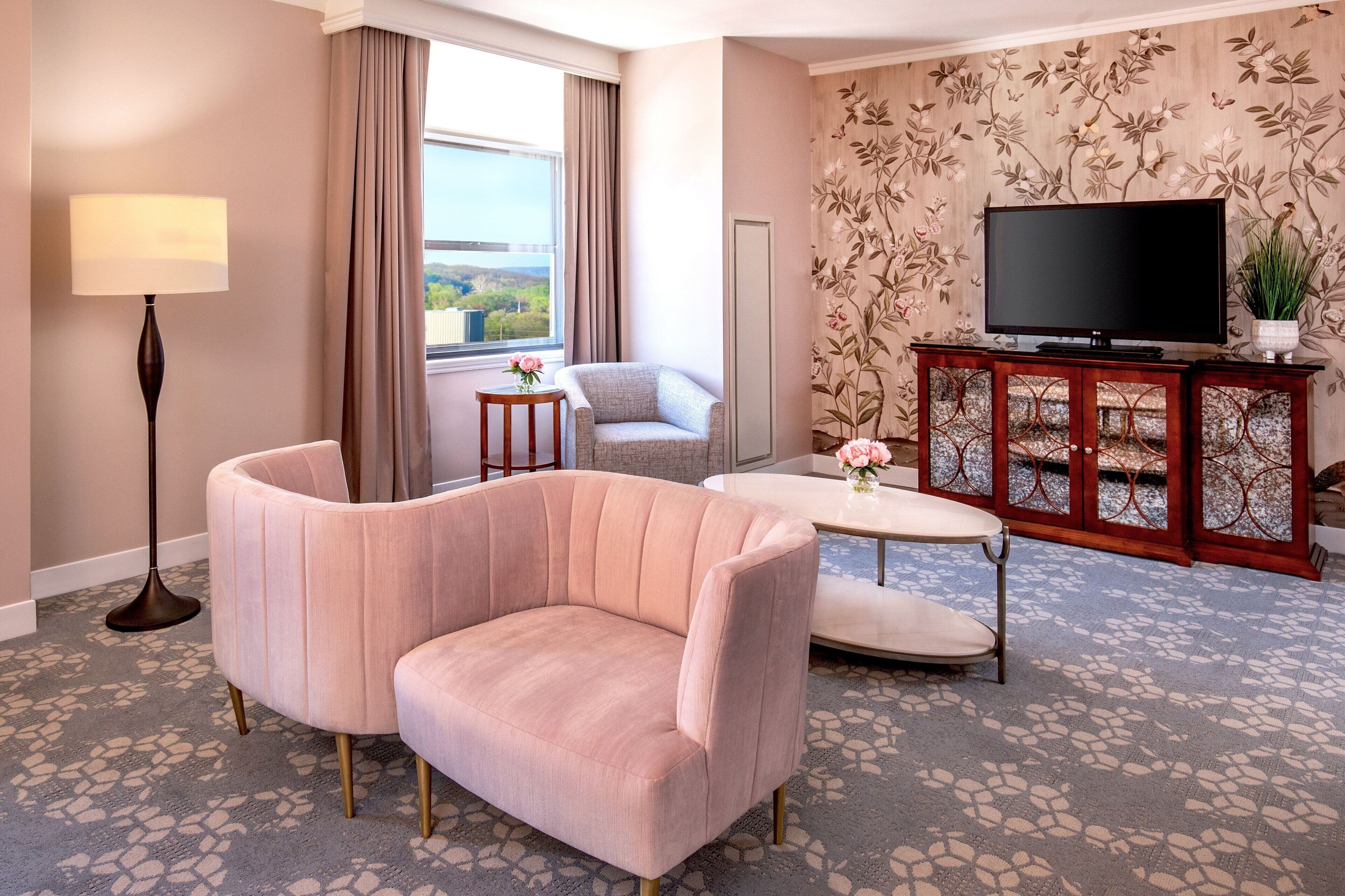 Une chambre d’hôtel avec un canapé rose, une petite table ronde, un téléviseur posé sur un meuble en bois, une lampe sur pied et un papier peint à motifs floraux.