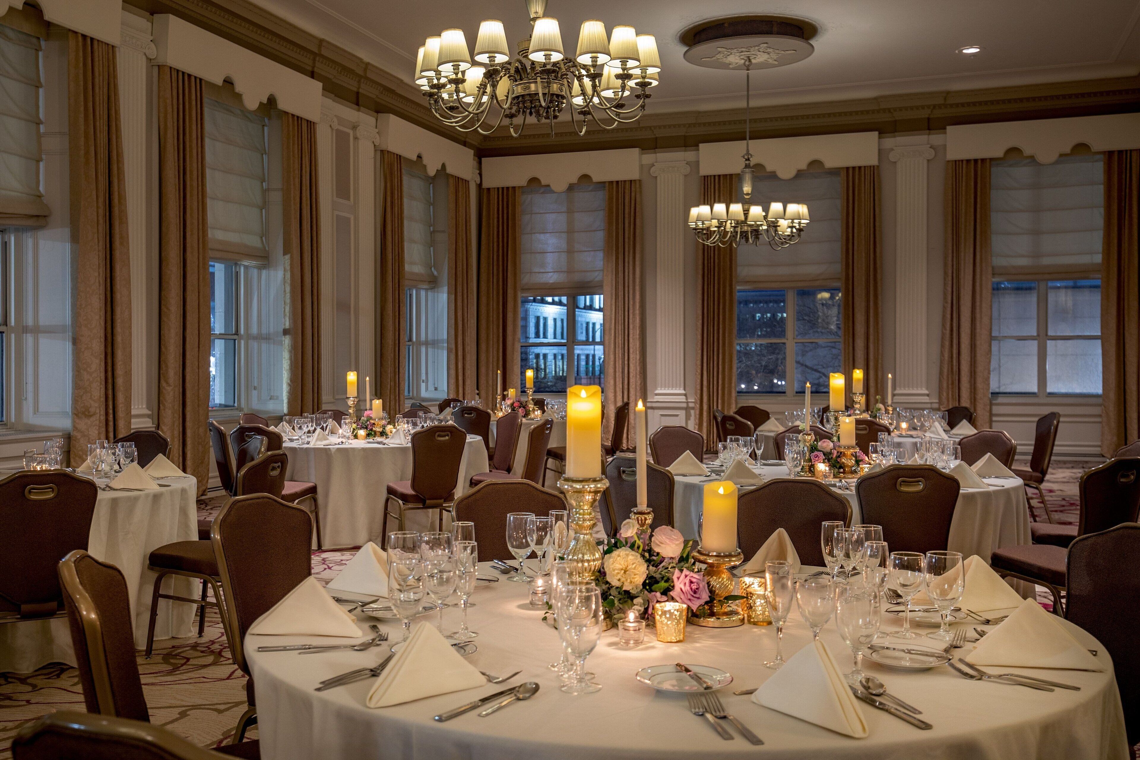 A banquet hall with round tables set for dining, chandeliers, and large windows with curtains.