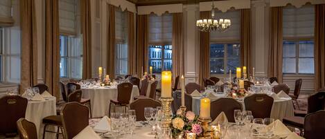 Un salón de banquetes con mesas redondas dispuestas para comer, candelabros y ventanas amplias con cortinas.