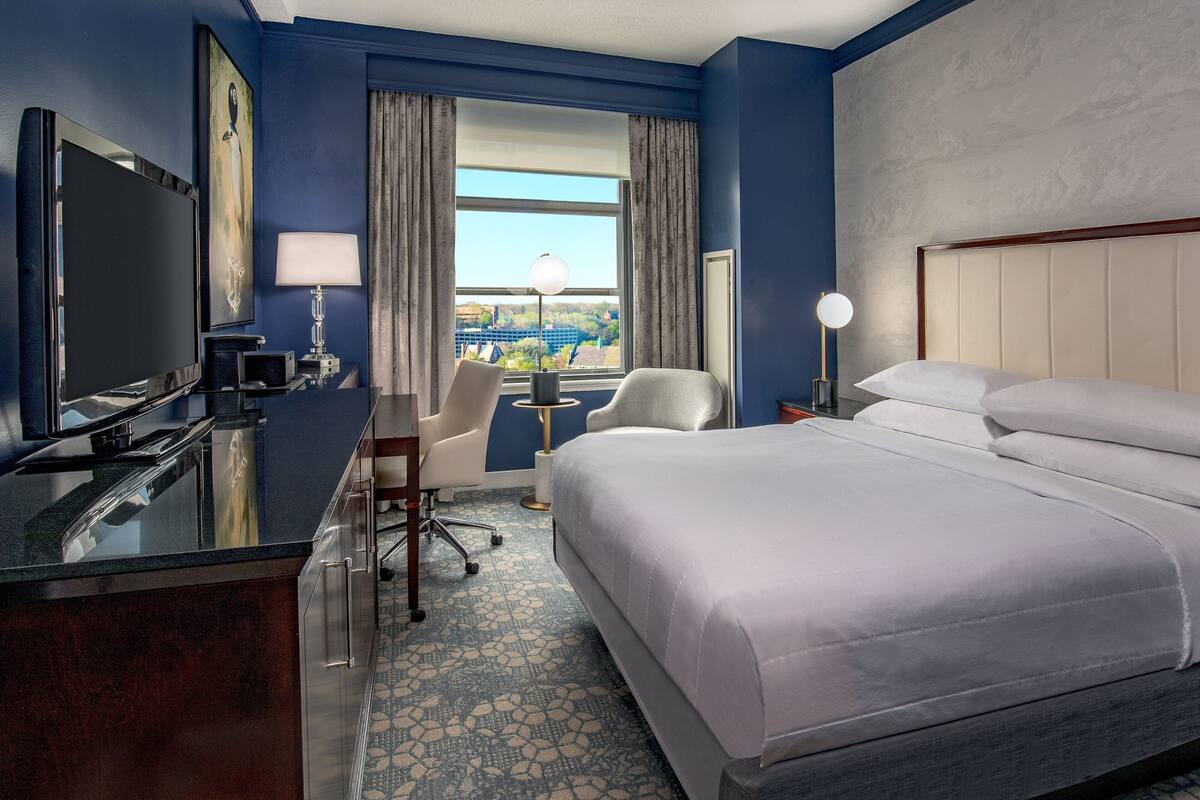 Een hotelkamer met een bed, bureau, televisie en uitzicht op de stad.