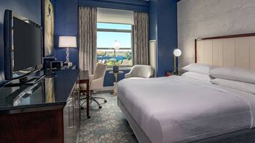 A hotel room with a bed, desk, TV, and a view of the city through the window.