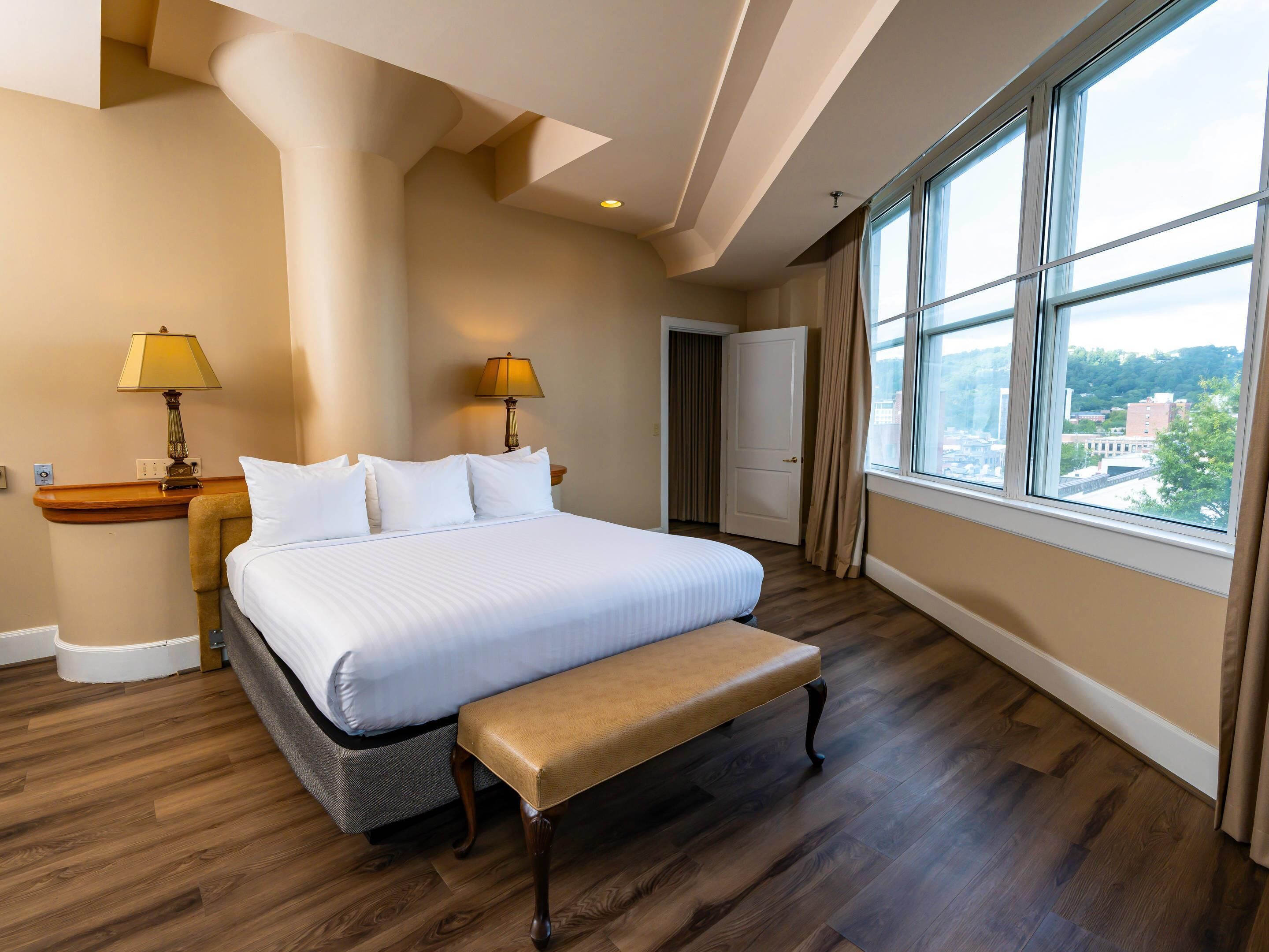 A hotel room with a large bed, two bedside lamps, a bench, a view of the city through large windows, and a door leading to another room.