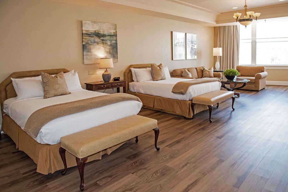 Une chambre d’hôtel avec deux lits, un banc, une petite table, une lampe et une fenêtre avec des rideaux.