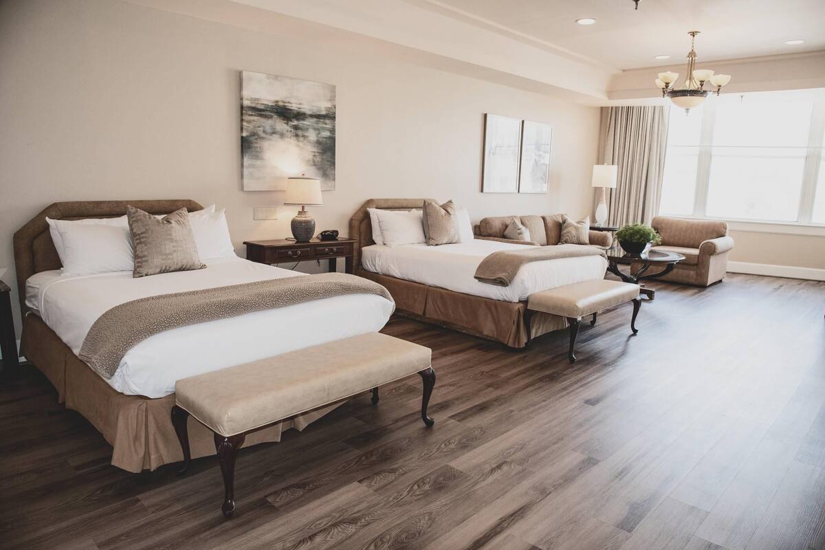 Une chambre d’hôtel avec deux lits, un banc, une chaise, une lampe et une fenêtre avec des rideaux.